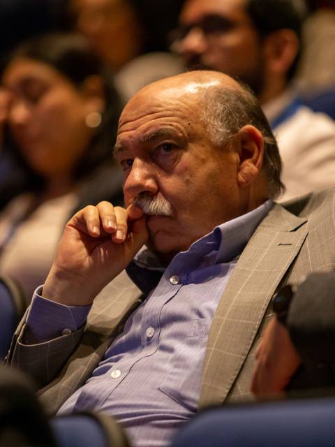 Un hombre con bigote se encuentra pensativo en un auditorio durante un evento.