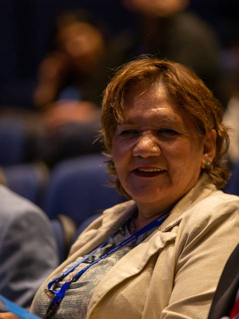 Una mujer sonriente con cabello rizado, sentada en una sala llena de personas.