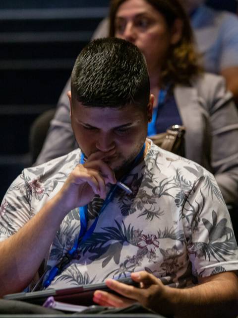 Un hombre con una camisa de flores está sentado en una sala de conferencias, concentrado en su dispositivo móvil.