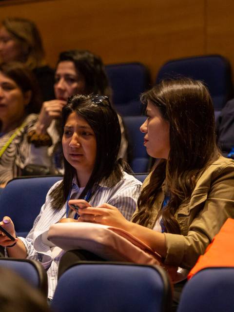 Un grupo de personas sentadas en un auditorio, algunas conversando y otras prestando atención.