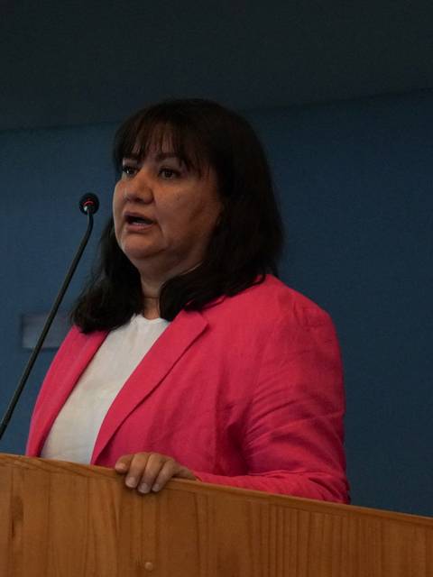 Una mujer con blusa blanca y chaqueta rosa habla en un podio de madera.