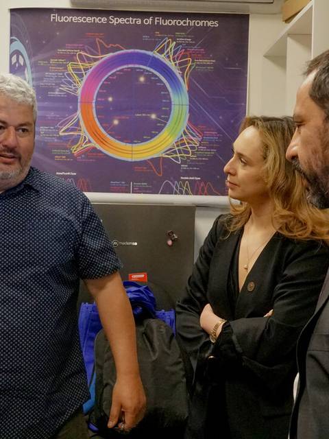 Tres personas conversan en un laboratorio con equipamiento científico y un gráfico en la pared.