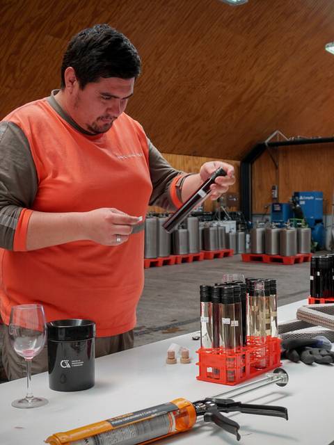 Un hombre trabaja en un taller, manipulando tubos de ensayo y herramientas de laboratorio mientras hay copas de vino en la mesa.