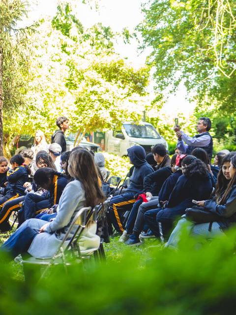 Un grupo de estudiantes sentados al aire libre, rodeados de vegetación, en un evento escolar.