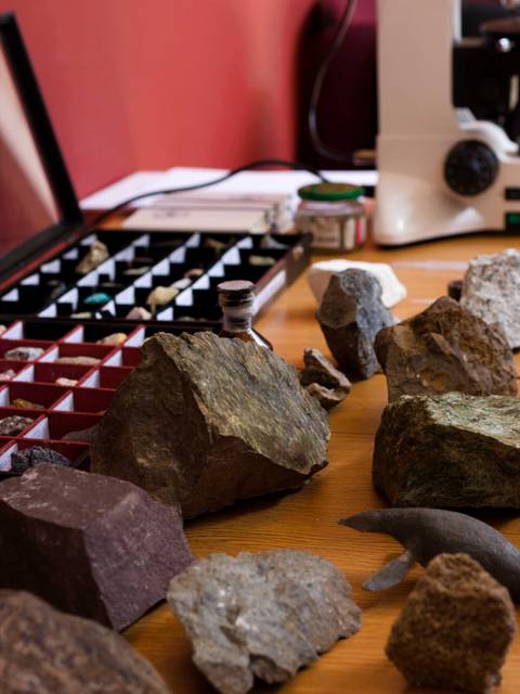 La imagen muestra una colección de rocas y minerales dispuestos sobre una mesa, junto a un microscopio.
