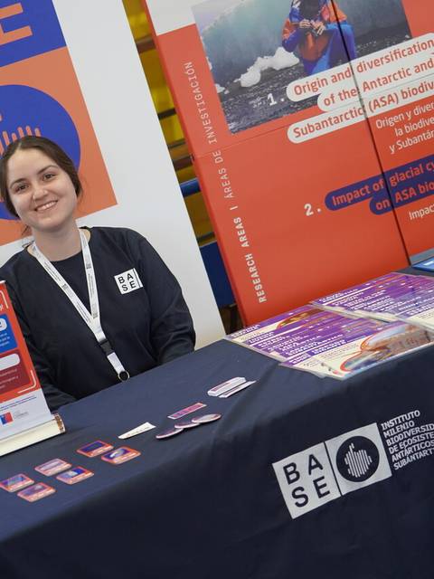 Una mujer sonriente está sentada detrás de una mesa con material informativo en un evento.