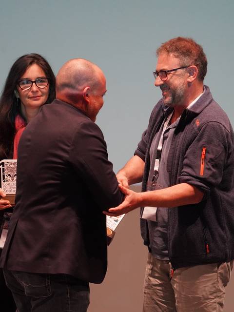 Tres personas interactuando en un evento, mientras uno de ellos recibe un premio.