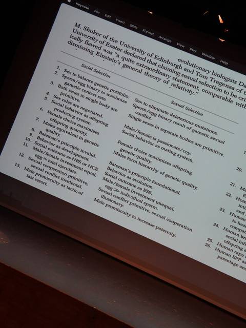 Un orador en un escenario presenta una diapositiva con texto sobre un tema académico.