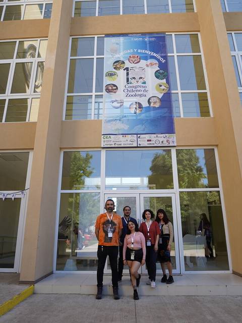 Un grupo de personas posando frente a un edificio con un cartel que presenta un evento.
