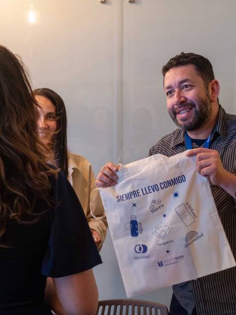 Un hombre sonriente muestra una bolsa de tela con un mensaje a un grupo de mujeres en un entorno de reunión.