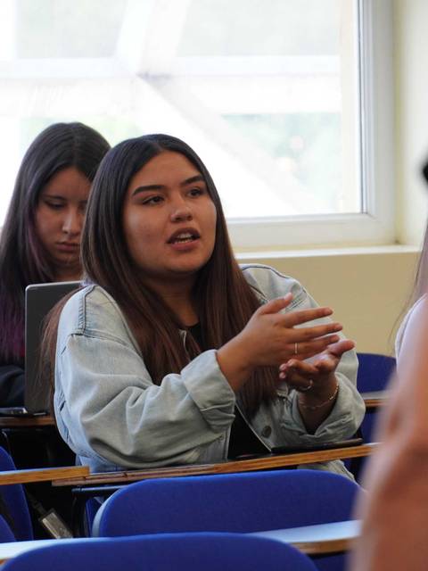 Una estudiante está participando activamente en una discusión en clase mientras otra persona la observa de fondo.