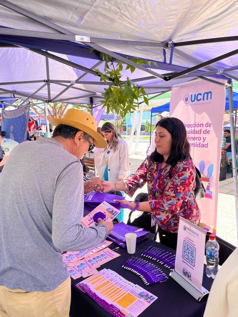 Una mujer entrega información a un hombre en un stand al aire libre, rodeado de folletos y una pancarta.