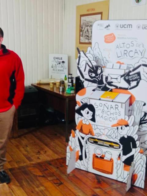 Un hombre de pie vestido con un suéter rojo junto a un panel informativo colorido en un ambiente interior.