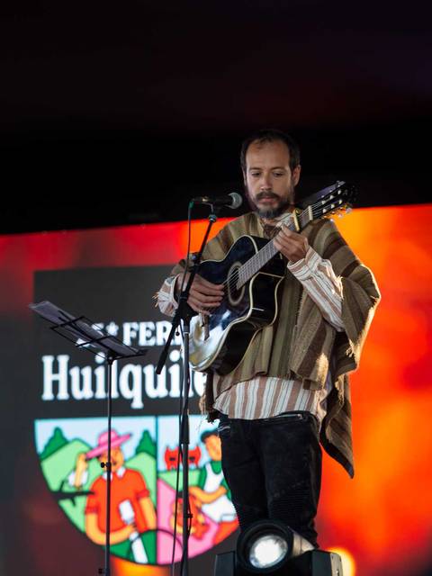 Un músico interpretando en un escenario durante un festival, vestido con un poncho y tocando la guitarra.