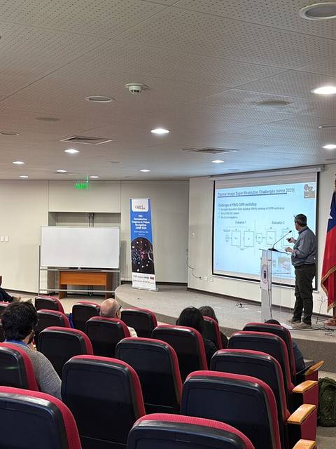 Una persona presenta en un aula con asientos vacíos y una pantalla de proyección.