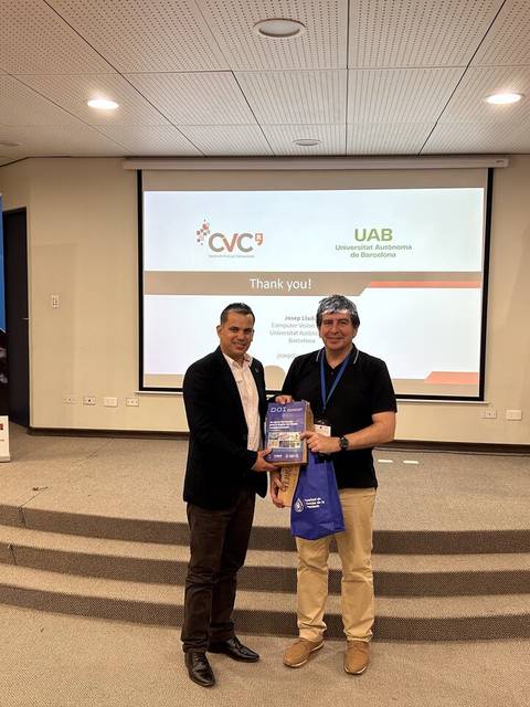 Dos personas posan juntas en un evento, una de ellas sostiene un premio, mientras que de fondo se observan pantallas y una bandera.