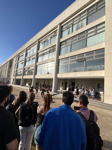 Un grupo de personas observa un evento frente a un edificio moderno durante un día soleado.
