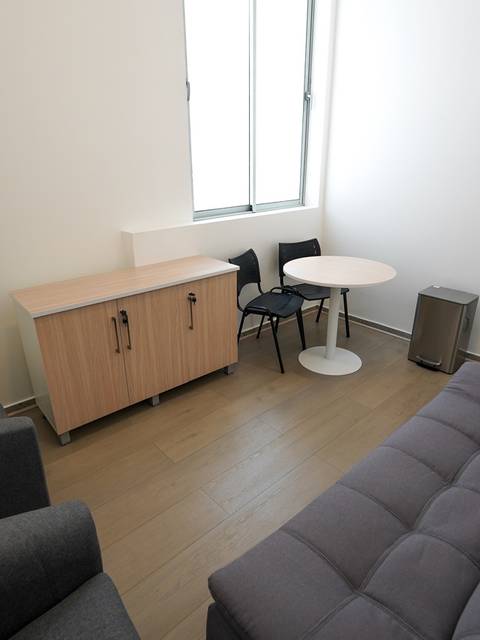 El espacio muestra una sala con un sofá gris, dos sillas, una mesa pequeña y un mueble de almacenamiento.