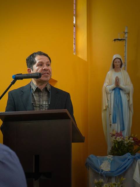 Un hombre hablando frente a un atril en un ambiente religioso con una estatua de la Virgen María al fondo.