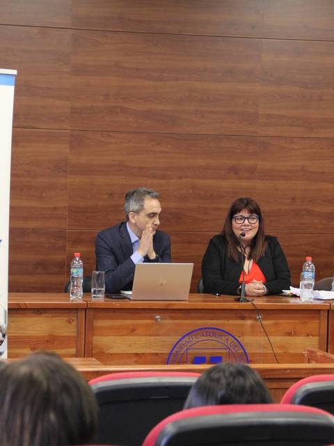 Una conferencia en una sala con un banner de la UCM y tres ponentes en una mesa.
