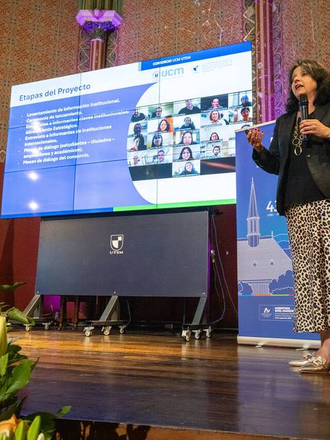 Una mujer presenta un proyecto frente a una pantalla durante un evento, mientras se conecta con otros participantes a través de videoconferencia.
