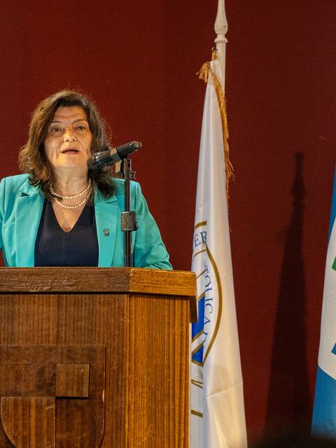 Una mujer está hablando en un podio durante un evento oficial, rodeada de banderas.