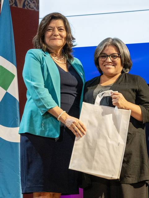 Dos mujeres sonrientes posan en un evento formal mientras sostienen una bolsa blanca.