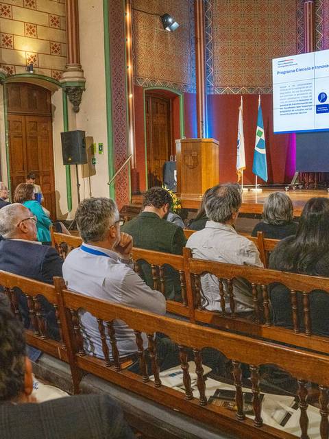Una conferencia con un ponente presentando información en una pantalla frente a una audiencia atenta.