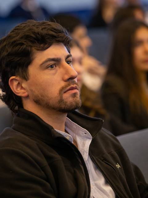 Un hombre sentado en un auditorio escucha atentamente mientras hay una multitud de personas detrás.