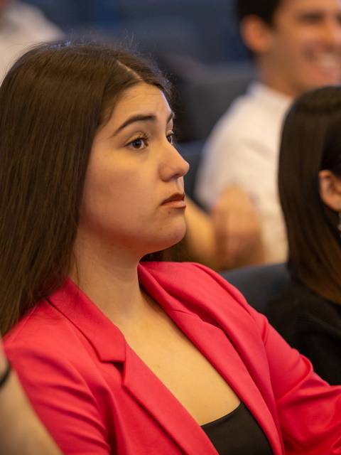 Una mujer joven con una chaqueta rosa escucha atentamente en un evento o conferencia.