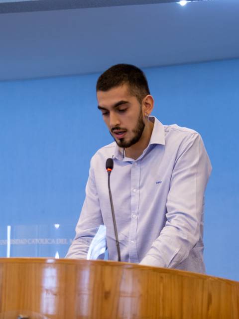 Un joven habla en un podio dentro de un auditorio con fondo azul.