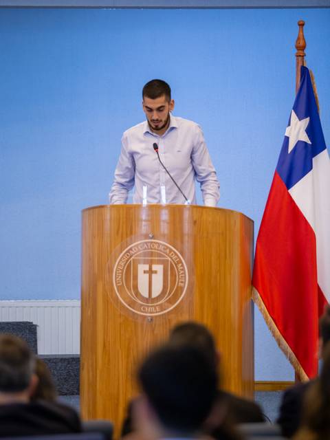 Un joven está hablando en un podio frente a una audiencia con una bandera chilena al fondo.