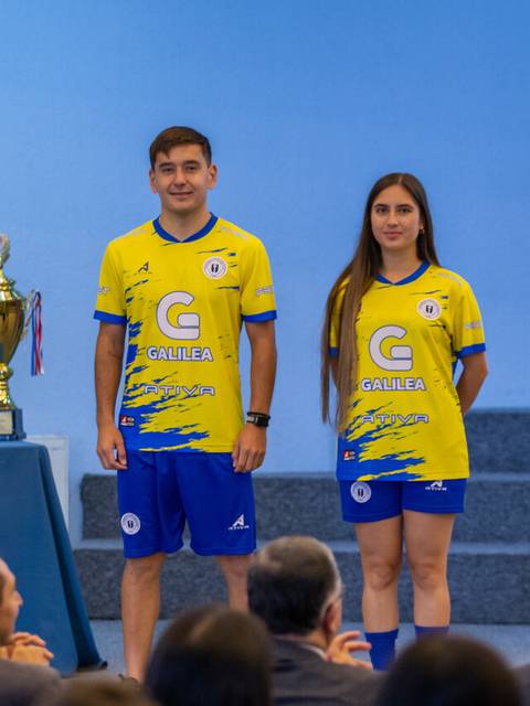 Dos jóvenes luciendo camisetas de un equipo de fútbol junto a trofeos en un evento.
