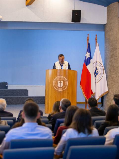 Un orador se dirige a un auditorio durante un evento en un salón con banderas.