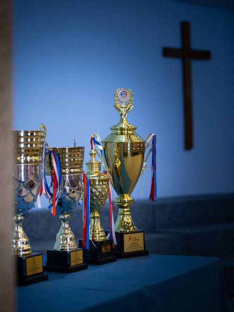 Una colección de trofeos dorados y plateados sobre una mesa con una cruz en la pared al fondo.