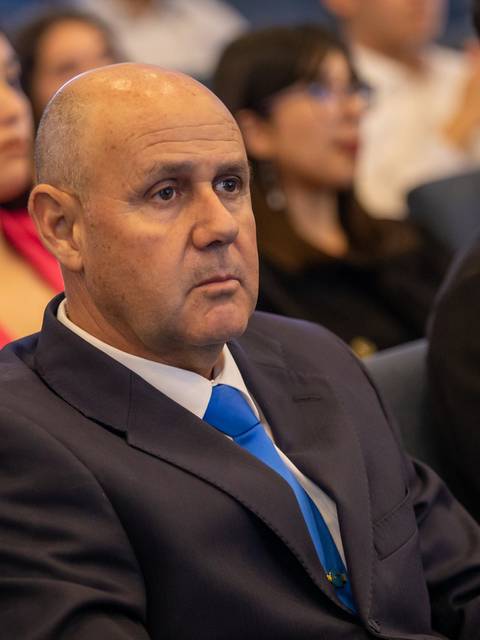 Un hombre de cabello corto y canoso, vestido con un traje oscuro y una corbata azul, escucha atentamente en un evento con un público diverso al fondo.