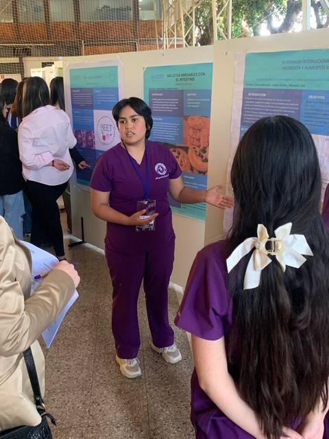 Una estudiante presenta su proyecto en una exposición frente a un grupo de personas interesadas.