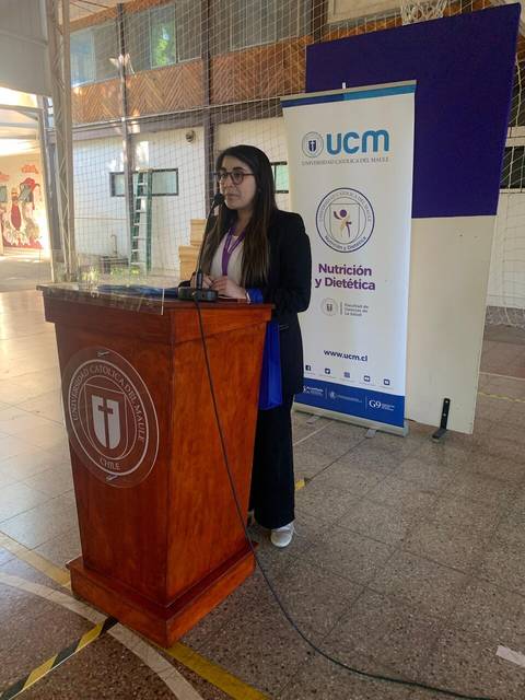 Una mujer está presentando en un evento, frente a un podio con un banner sobre nutrición y dietética.