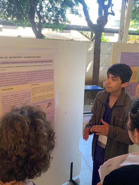 Un joven presenta un póster en un evento académico ante un grupo de personas interesadas.