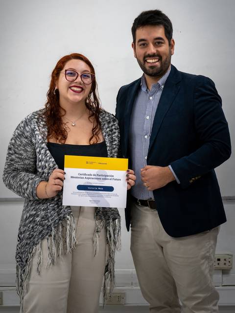 Una mujer y un hombre posan juntos sosteniendo un certificado en un aula.