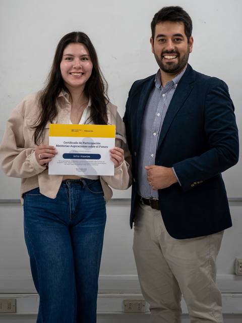Una joven sostiene un certificado mientras posa junto a un hombre en un aula.