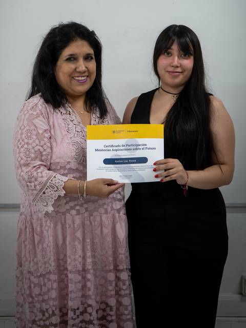 Dos mujeres posan juntas sosteniendo un certificado en un entorno de aula.