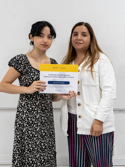 Dos mujeres posan juntas sosteniendo un certificado en un entorno educativo.