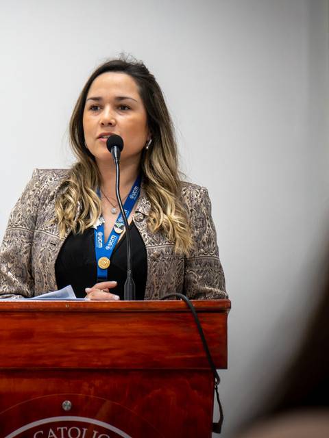 Una mujer habla en un podio durante un evento, luciendo una chaqueta brillante y sosteniendo un micrófono.