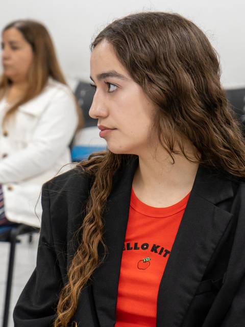Una joven con chaqueta negra y camiseta roja de Hello Kitty escucha atentamente en un evento con otras personas al fondo.
