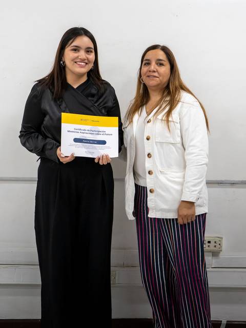 Dos mujeres posan sonrientes, una de ellas sostiene un certificado en sus manos.