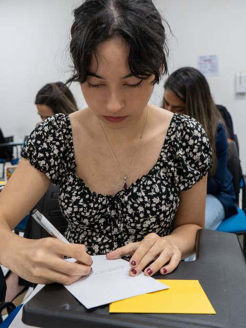 Una joven con un vestido de flores escribe en una hoja sobre una mesa en un aula.