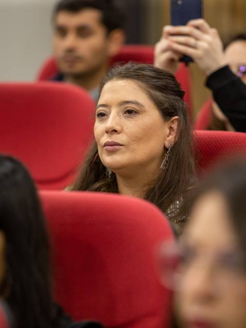 Una audiencia sentada en sillas rojas, con un enfoque en una mujer al frente que parece atenta.
