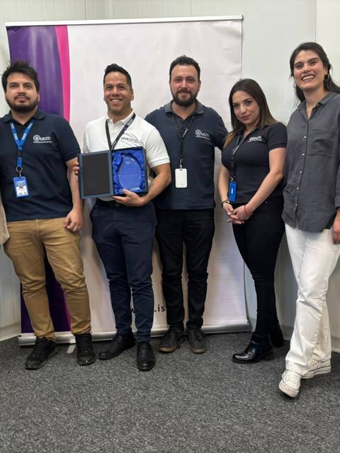 Un grupo de seis personas sonríen mientras sostienen un reconocimiento en un ambiente académico.