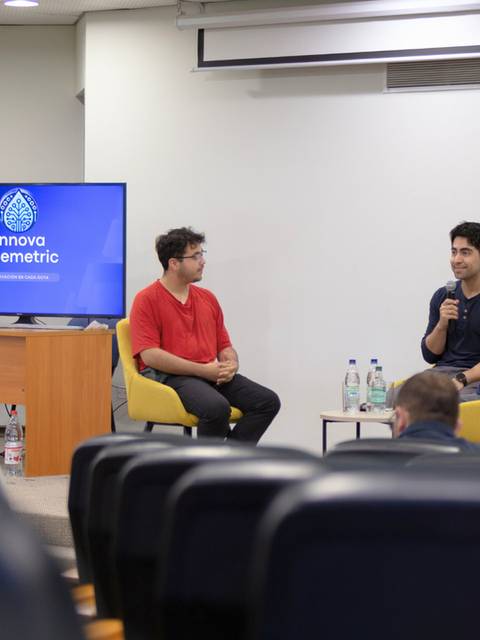 Un panel de discusión en un evento, con tres participantes en un escenario y una pantalla mostrando el logo de una empresa.